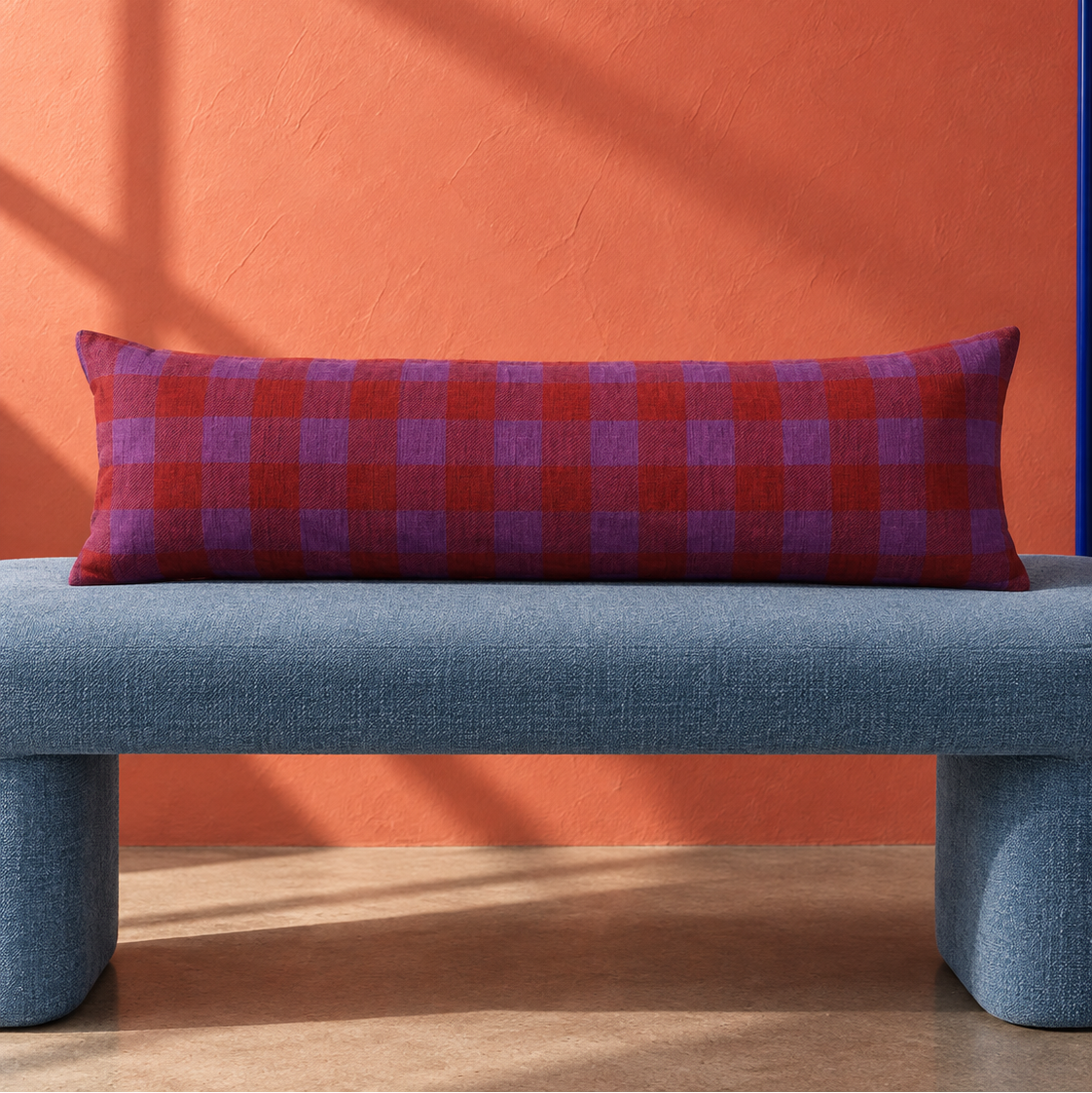 A long rectangular throw pillow with a red and purple plaid pattern, resting on a blue upholstered bench against a textured orange wall.