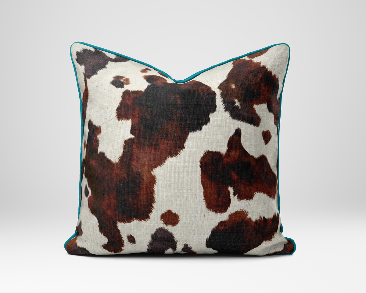 A square throw pillow featuring a brown and black cowhide pattern on an off-white background, finished with a thin teal piped edge, shown against a neutral light gray background.