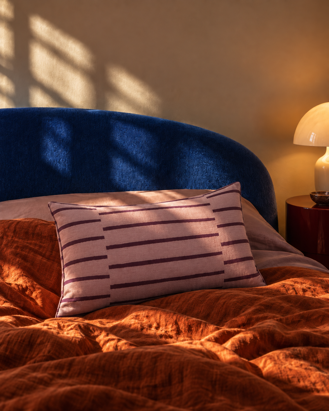 A striped lumbar pillow in a dusty pink fabric with dark purple stripes, styled on a bed with a blue headboard, rust-orange textured bedding, and warm sunlight casting shadows.