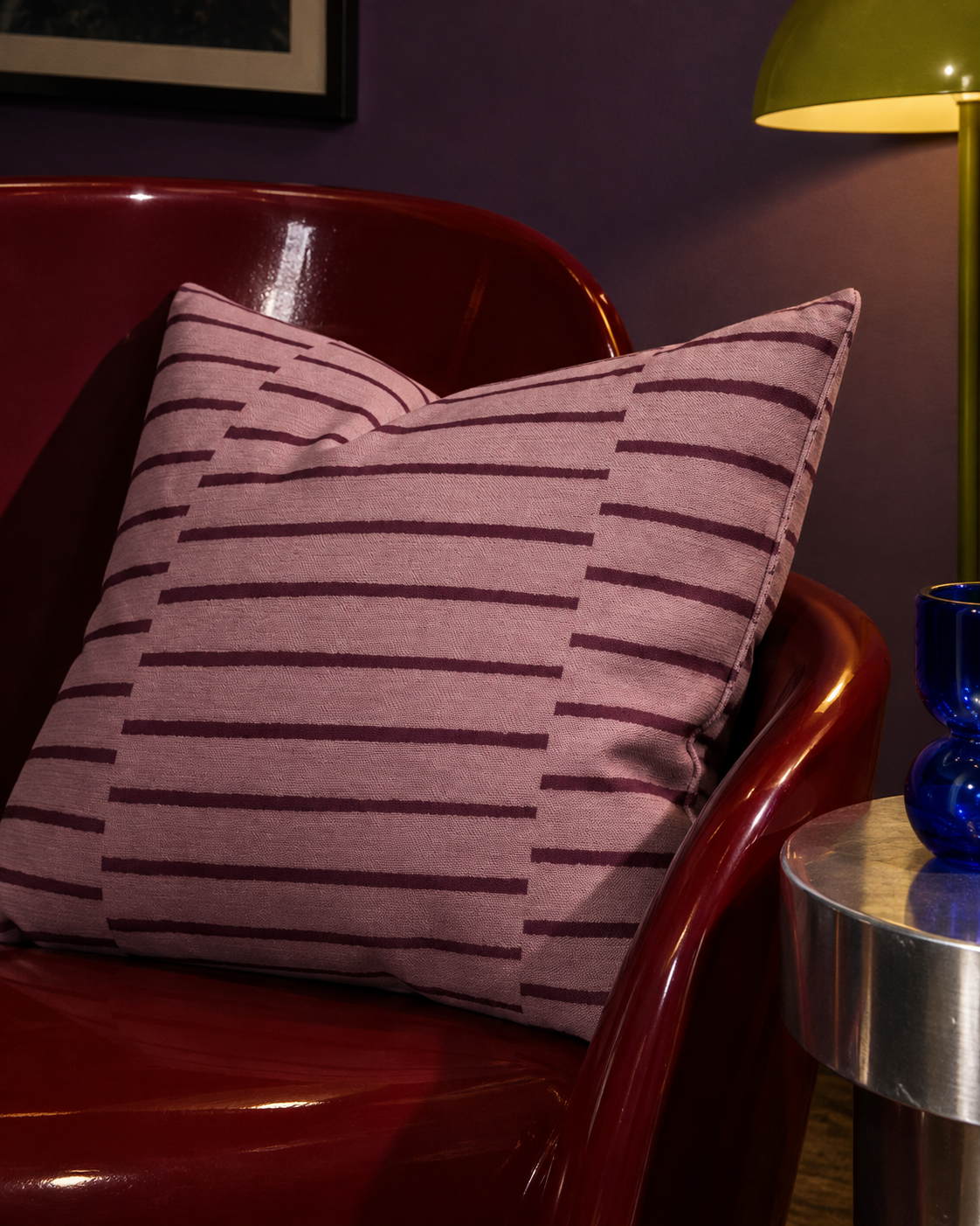 A square throw pillow with a dusty pink and oxblood striped pattern, resting on a glossy dark red armchair in a dimly lit room.