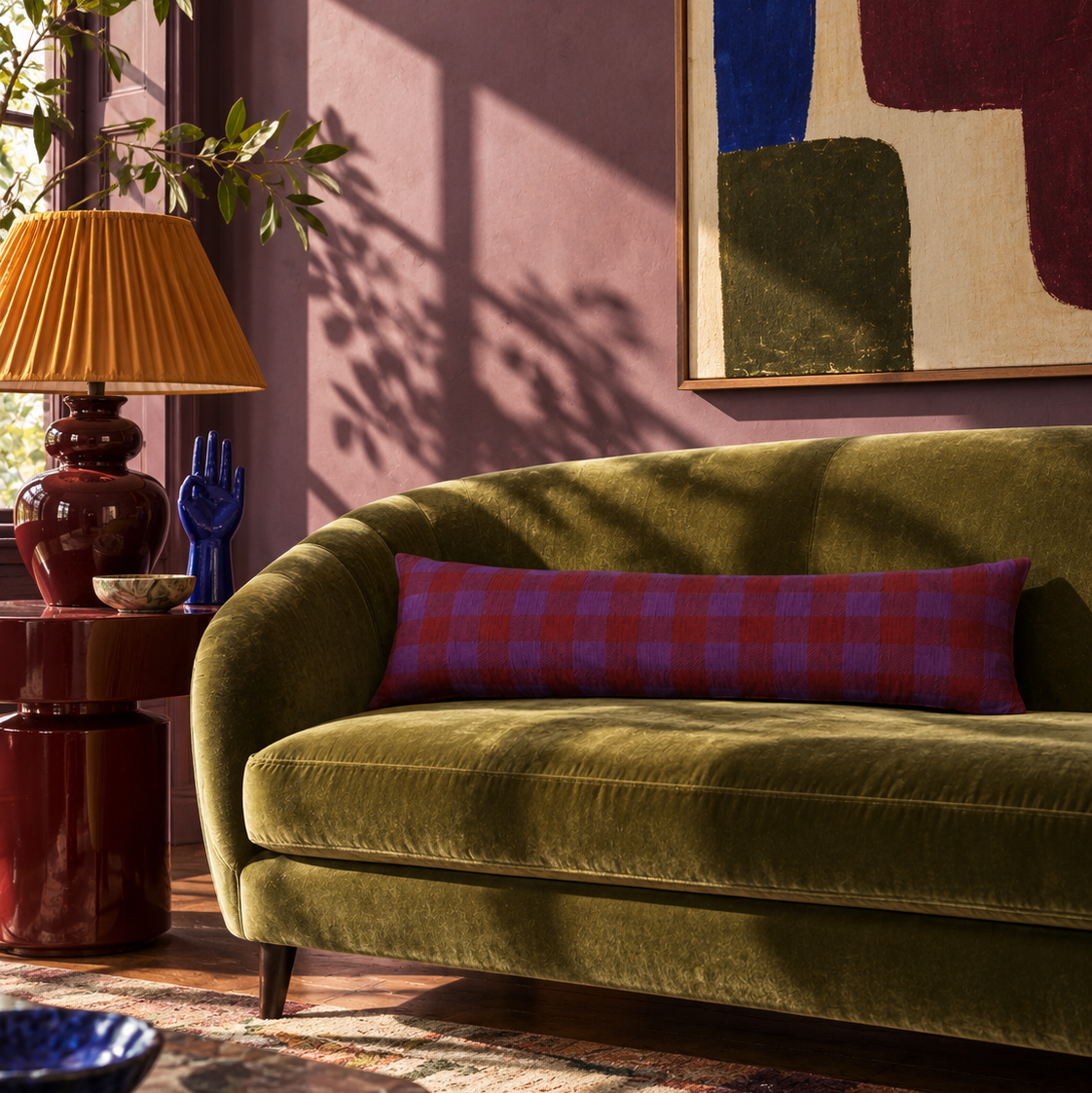 An olive green velvet sofa in a styled living room setting, featuring a red and purple plaid lumbar pillow, a warm orange lamp, and a patterned rug.